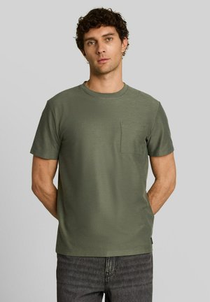 Junger Mann mit lockigem dunklem Haar, der ein olivgrünes T-Shirt mit kurzen Ärmeln und Brusttasche und dunkelgraue Jeans vor einem einfarbigen Hintergrund trägt.