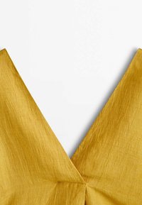 Gros plan sur un tissu jaune doré avec un décolleté en V profond et une texture visible sur fond blanc.