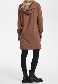 Tunica cappuccio marrone con maniche lunghe, caratterizzata da un taglio dritto e dettagli a contrasto, abbinata a pantaloni neri slim-fit e stivaletti neri alla caviglia.