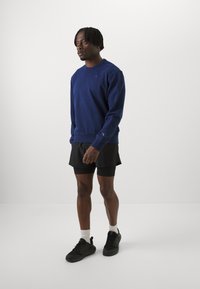 Felpa navy con polsini e orlo a coste, caratterizzata da un piccolo logo. Abbinata a pantaloni corti neri e sneakers nere. Texture del tessuto liscia.