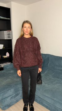Weinroter strukturierter Pullover mit Rundhalsausschnitt, kombiniert mit schwarzen Hosen mit geradem Bein und schwarzen Slippern. Hält eine kleine schwarze Tasche.