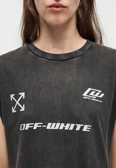 Κοντινό πλάνο ατόμου που φορά ξεθωριασμένη μαύρη αμάνικη μπλούζα με λευκό κείμενο και λογότυπα "OFF-WHITE" τυπωμένα στο στήθος.