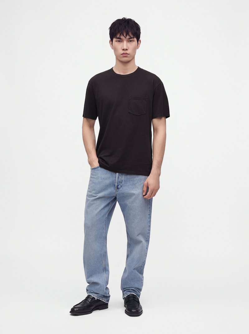 Jeune homme aux cheveux noirs portant un t-shirt noir, un jean bleu clair et des chaussures noires, debout avec une main dans la poche sur un fond uni.