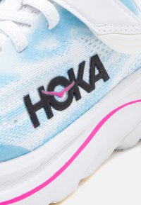 Εγγύς λήψη αθλη τικού παπουτσιού Hoka σε ανοιχτό μπλε και λευκό χρώμα με μαύρο λογότυπο "HOKA" και ροζ τόνους στη σόλα και το σημάδι ελέγχου του λογότυπου.
