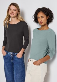 Dos mujeres están de pie una al lado de la otra, vistiendo camisetas de manga larga: una de color gris oscuro y la otra de un tono aguamarina claro. Ambas llevan jeans casuales y un corte relajado.