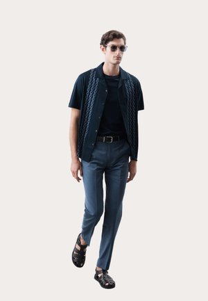 Camisa de manga corta azul marino con acentos estampados, camiseta negra y pantalones gris ajustados. Sandalias negras de tiras complementan el look.