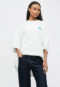 Νέα γυναίκα με σγουρά μαλλιά που φοράει ένα φαρδύ λευκό t-shirt με μπλε εκτύπωση "American Vintage" και σκούρα μπλε τζιν, ποζάροντας με το ένα χέρι στην τσέπη.