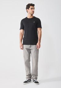 Schwarzes Baumwoll-T-Shirt mit kurzen Ärmeln und einem kleinen wei�ßen Logo auf der Brust, kombiniert mit hellgrauen Jeans und schwarzen Sneakers.