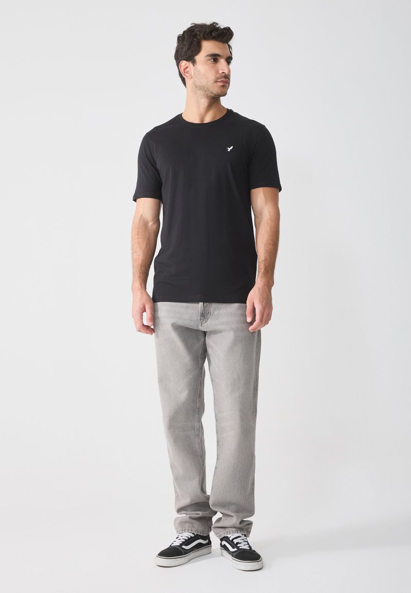 Schwarzes Baumwoll-T-Shirt mit kurzen Ärmeln und einem kleinen weißen Logo auf der Brust, kombiniert mit hellgrauen Jeans und schwarzen Sneakers.