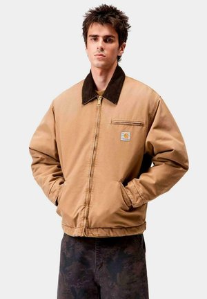 Chaqueta de algodón marrón con un cuello oscuro, cierre con cremallera y un bolsillo frontal. Llevada sobre un suéter texturizado amarillo. Se ven pantalones oscuros.