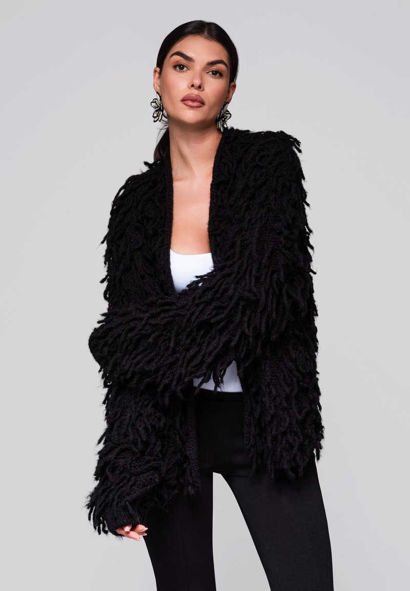 Svart fluffig cardigan med ett texturerat, lurvigt stickmönster. Långa ärmar och öppen framstil. Bärs över en vit linne och svarta byxor.