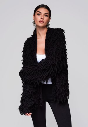 Cardigan noir en laine shaggy avec un design tricoté texturé et duveteux. Manches longues et style à ouverture frontale. Porté par-dessus un débardeur blanc et un pantalon noir.