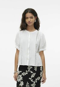 Valge lühikeste varrukatega blouse, millel on ümmargune krae, esipaneelil nööbid ja pitsaktsentid. Must seelik, millel on beež lilleline tikand.