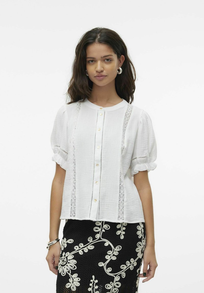 Valge lühikeste varrukatega blouse, millel on ümmargune krae, esipaneelil nööbid ja pitsaktsentid. Must seelik, millel on beež lilleline tikand.