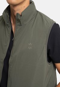 Veste sans manches verte avec col montant, dotée d'une fermeture éclair à l'avant et d'un logo discret. Le tissu semble léger avec une texture lisse.