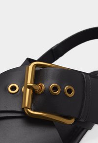 Κοντινό πλάνο μαύρης δερμάτινης ζώνης με χρυσή μεταλλική αγκράφα και ταιριαστά χρυσά μεταλλικά τρύπες σε απλό φόντο.