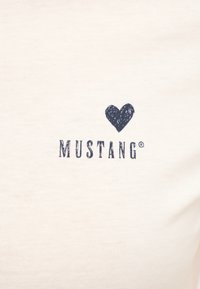 Camiseta de algodón crema que presenta un corazón azul y el texto 'MUSTANG' debajo en una fuente casual. Textura suave con un corte clásico.