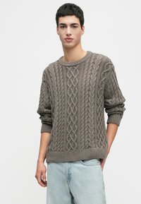 Junger Mann, der einen taupefarbenen, grob strukturierten Pullover mit hochgeschobenen Ärmeln und hellblauen Jeans trägt und vor einem schlichten weißen Hintergrund steht.