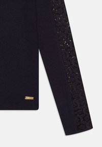Haut à manches longues bleu marine foncé avec texture côtelée, présentant des détails en dentelle sur la manche gauche et une petite étiquette de marque dorée en bas.