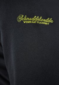 Tessuto nero con una trama testurizzata, caratterizzato da testo ricamato verde che recita "Schmuddelvedda WENN DAS PLADDBERT."