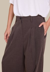 Brune høytlivsbukser med legg, laget av strukturert stoff. Har knappelukking og sidelommer. Kombinert med en hvit topp.