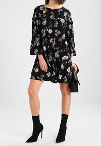 Czarna sukienka w kwiaty z fioletowymi i białymi motywami, z rękawami o długości trzy czwarte, marszczonym fasonem, zestawiona z czarnymi botkami do kostek i kopertówką.