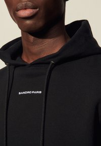 Sudadera de algodón negra con cordones, con "SANDRO PARIS" bordado en blanco en el pecho, textura suave y corte holgado.
