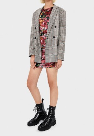 Donna che indossa un mini abito in velluto floreale, blazer doppiopetto a quadri e stivali neri stringati da combattimento, in piedi davanti a uno sfondo semplice.