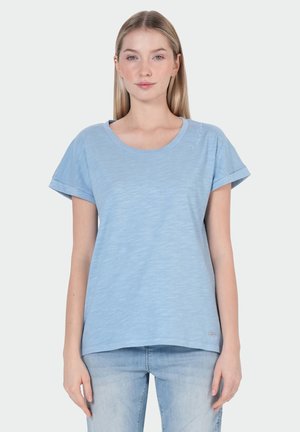 Jonge vrouw staat, draagt een lichtblauw T-shirt met korte mouwen en lichtgewassen spijkerbroek tegen een effen achtergrond.