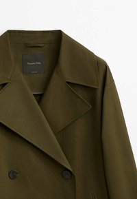 Manteau double boutonnage vert olive avec grands revers, boutons noirs et texture lisse. Étiquette visible à l'intérieur du col.