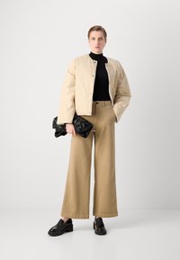 Beige kort jakke, svart genser med høy hals, brune vide bukser og svarte platå lofer. Holder en svart teksturert veske.