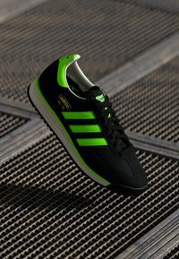 Black athletic shoe with green accents, featuring three stripes, a textured upper, rubber sole, and branding in gold on the side.