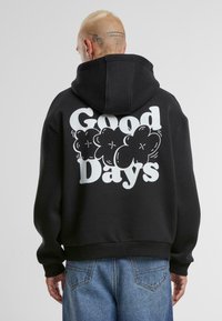 Zwarte hoodie met een grote witte grafiek met de tekst "Good Days" omgeven door wolkvormen. Zachte textuur en een relaxte pasvorm.