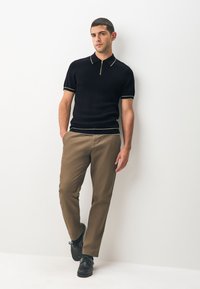 Zwart geribd poloshirt met gouden biezen en een ritssluiting bij de kraag, gecombineerd met beige broek en zwarte schoenen. Eenvoudig, modern ontwerp en pasvorm.
