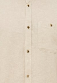 Μπεζ πουκάμισο με κουμπιά σε καφέ χρώμα και τσέπη στο στήθος, κατασκευασμένο από υφάσμα με υφή.