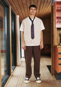 Gilet beige lavorato a maglia sopra una t-shirt bianca, cravatta a righe blu navy e viola, pantaloni marroni e sneaker bianche e nere in un interno di legno.