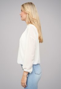 Hvid bluse med gennemskinnelige lange ærmer, afslappet pasform og rund halsudskæring, parret med lyseblå tætsiddende jeans. Glat tekstur, minimal detaljering.