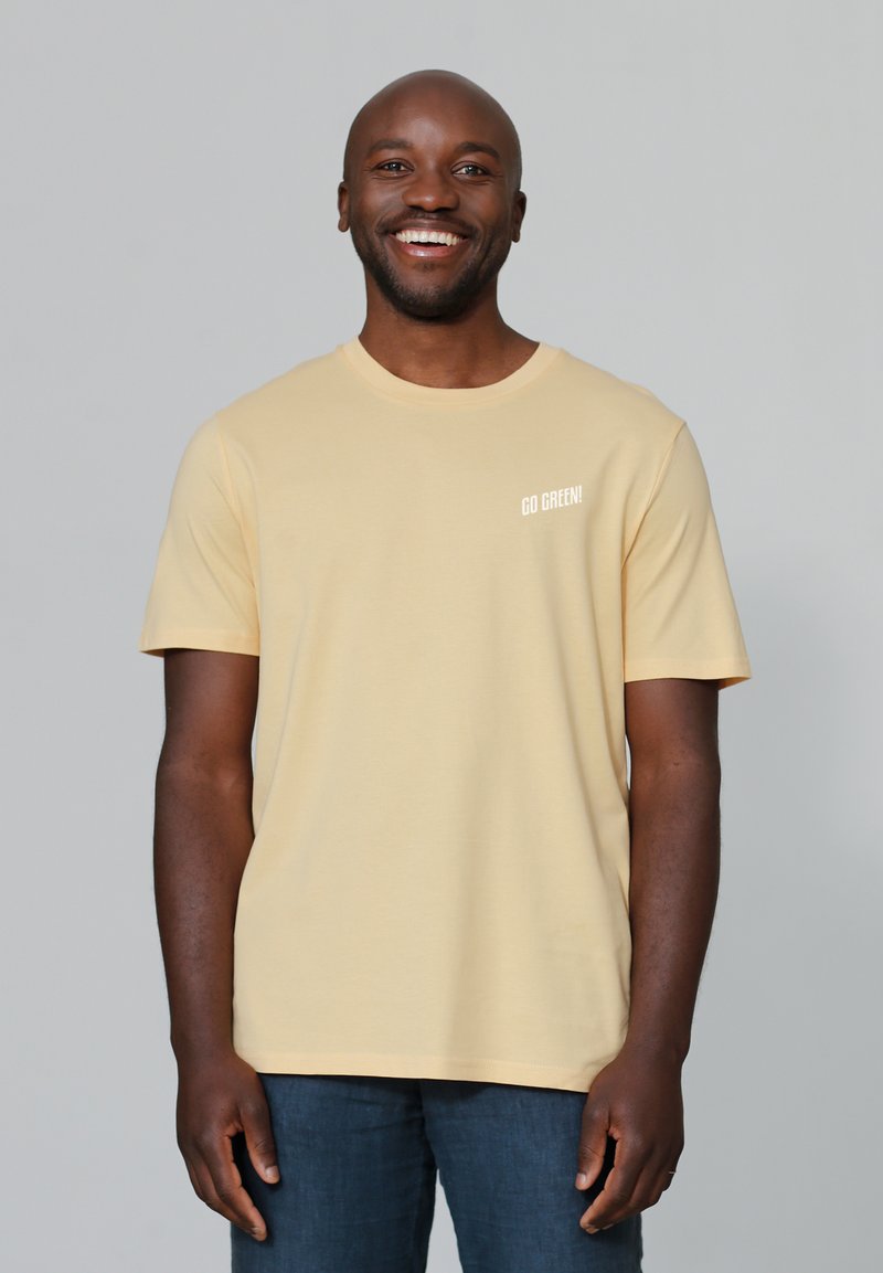Helles gelbes T-Shirt mit kurzen Ärmeln, das den Text "GO GREEN!" in Weiß auf der Brust trägt. Der Stoff scheint weich und glatt zu sein; das Modell steht vor einem neutralen Hintergrund.