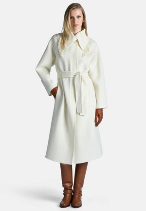 Femme portant un long manteau blanc ceinturé avec des détails de col à franges, des gants en cuir marron et des bottes hautes marron assorties, debout avec une main dans la poche.