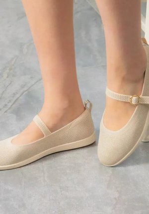 Pieds portant des ballerines Mary Jane en maille beige avec une petite boucle sur la bride, debout sur un sol en marbre gris.