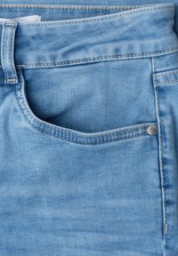 Jean denim bleu clair en gros plan montrant la poche avant, la passants de ceinture et un rivet argenté sur la texture du tissu.