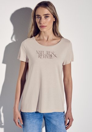 T-shirt corto a maniche corte color beige chiaro con scollatura a barca, con la scritta "NATURE & REVIVAL" stampata in marrone scuro. Tessuto morbido, vestibilità comoda.
