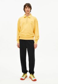 Gele trui met een halve rits en geborduurd detail. Gecombineerd met zwarte broek en gele sportschoenen met rode accenten.