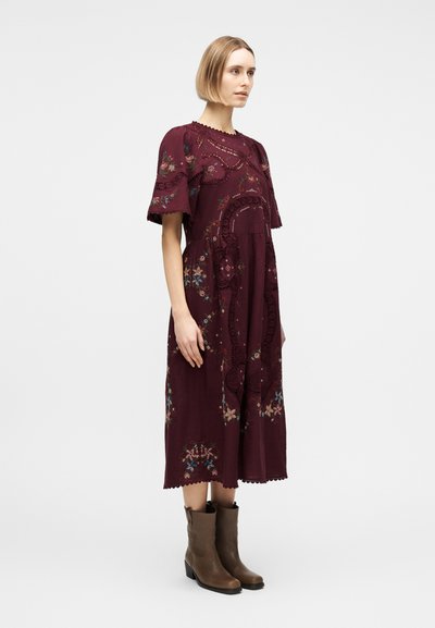 Γυναίκα που φοράει βουργουνδί midi φόρεμα με κεντήματα από λουλούδια και λεπτομέρειες από δαντέλα με ανοίγματα, συνδυασμένο με καφέ μποτάκια αστραγάλου, στέκεται στο προφίλ.