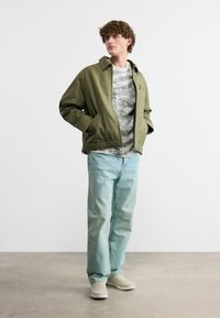 Νέος άνδρας που φοράει χακί μπουφάν, γκρι πουκάμισο με σχέδια, ανοιχτό μπλε τζιν και λευκά αθλητικά παπούτσια, στέκεται με τα χέρια στις τσέπες.