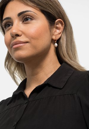 Gullfargede hoopøreringer med en dråpeformet hvit perleakcent; modellen har på seg en svart skjorte med knapper, med krage og myk stofftekstur.