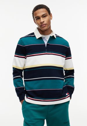 Młody mężczyzna ubrany w koszulę rugby z długim rękawem w paski: turkusowe, granatowe, białe i żółte, z białym kołnierzem oraz zielone spodnie, stojący z rękami w kieszeniach.