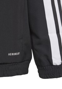 Sort atletisk jakke med hvide striber, elastiske manchetter og "AEROREADY" branding på kanten, lavet af et glat og let materiale.