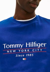 Koninklijk blauwe katoenen T-shirt met het "Tommy Hilfiger" logo in witte en rode tekst, met een kleine vlagafbeelding onderaan. Ronde hals ontwerp.