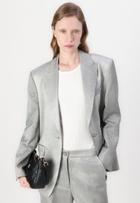 Lys grå todelt dress med en strukturert blazer og glatt tekstur, kombinert med en hvit ribbet topp og liten svart veske.